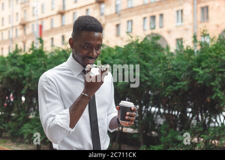 African american businessman in a rush. Man with takeaway coffee speaks on a speakerphone or records a voice message. Stock Photo