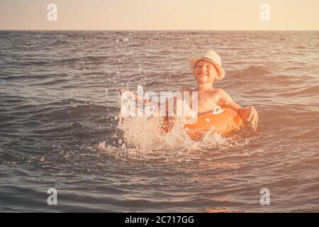 Cheerful Caucasian boy in a white hat on an orange swimming circle splashes his legs in the sea and smile. A child swims and has fun at the resort at