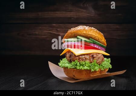 Classic burger made of bun with sesame seeds, beef pattie, lettuce, cheese, onions, cucumber, tomato on a sheet of baking paper on a wooden rustic bac Stock Photo