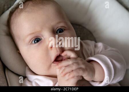 A close-up portrait of a surprised baby girl who opened her eyes wide and holds her hand in her mouth. three-month-old baby girl Stock Photo