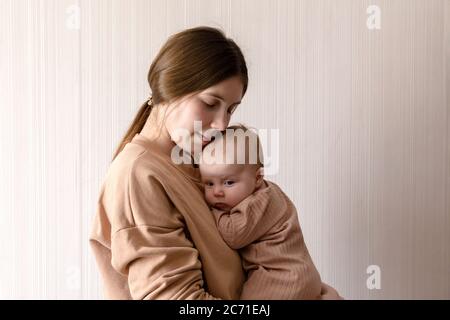 Cheerful beautiful young woman holding baby girl in her hands and looking at her with love at home