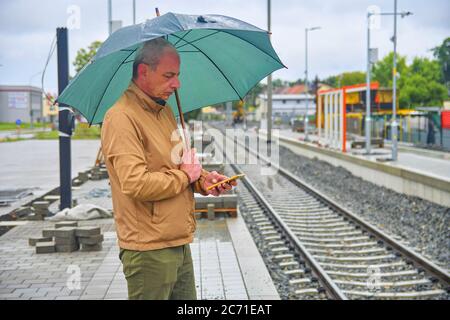 Portrait of a mature man texting on smartphone with an umbrella standing on the platform of a train station. Unfinished railway line. When the train Stock Photo