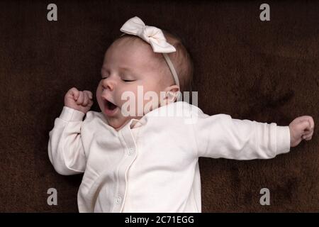 Newborn baby girl boy wants to sleep, rubs his eyes with his hand and yawns on white sheets from above. Sleep. Stock Photo