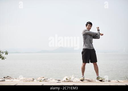 young asian adult man stretching arms outdoors by the sea