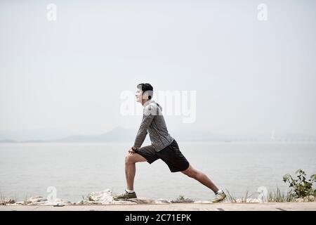 young asian adult man stretching legs outdoors by the sea