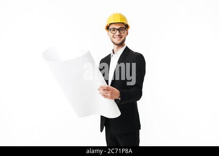Young bussinesman in eyeglasses and yellow safety helmet happily looking in camera holding sketch in hands over white background isolated Stock Photo