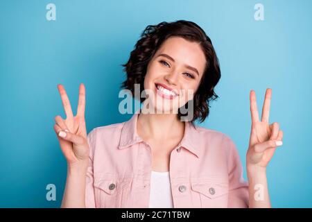 Photo of attractive cheerful lady short black hairdo raise hands showing v-sign symbols positive good mood saying hi to friends wear casual pink denim Stock Photo
