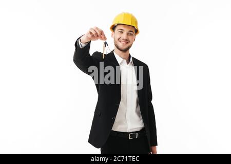 Young smiling bussinesman in yellow safety helmet happily looking in camera while holding keys of new flat in hand over white background Stock Photo
