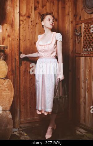 Young, good-humoured, red-blonde woman in dirndl with bag at the entrance of a mountain hut Stock Photo