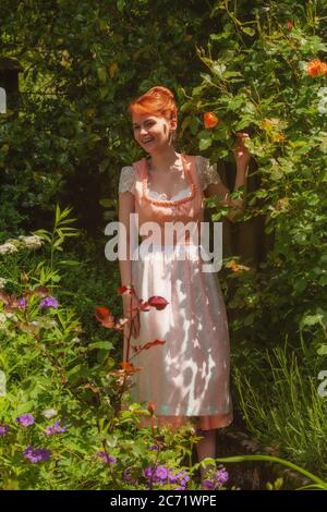 Young woman in dirndl laughing in your garden and happily holding a rose in her hand. Stock Photo