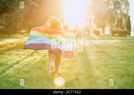 Back view of incognito young woman swinging rainbow flag on wind behind back. Unrecognizable girl running in park, enjoying beautiful summer sunset. Concept of lgbt, minorities tolerance. Stock Photo