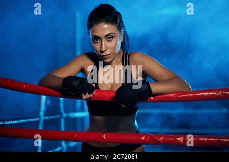Professional female kickboxer with strong face posing in smoky blue atmosphere, wearing bandages on hands. Fighter having rest after training, leaning on rope and looking at camera. Concept of boxing. Stock Photo