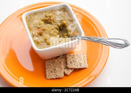 Split pea soup and crackers with a spoon in the square white bowl on an orange plate.  Homemade steaming hot soup healthy food for lunch or dinner. Stock Photo