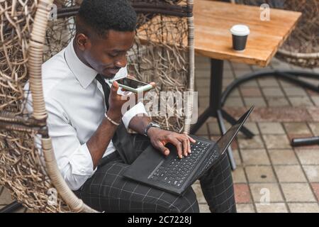 Business african american man holding smartphone, dictating voice message, recording audio, using voice mic. People works outdoors Stock Photo