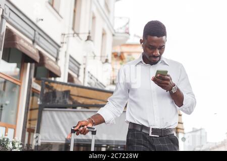 Young handsome black man with luggage using smartphone and listening music with earphones in the city Stock Photo