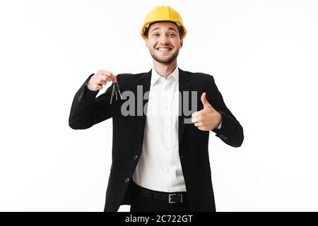 Young cheerful realtor in yellow safety helmet happily showing thumb up looking in camera with keys of new flat in hand over white background Stock Photo