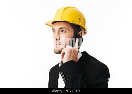 Young serious bussinesman in yellow safety helmet thoughtfully looking aside talking on cellphone over white background Stock Photo