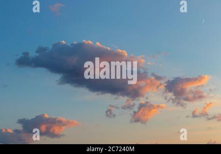Fluffy Cumulus clouds in blue sky at sunset with golden light and crescent moon, evening time Stock Photo