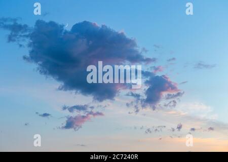 Soft pink purple fluffy Cumulus clouds in blue sky as sun begins to set at sunset with golden light and crescent moon Stock Photo