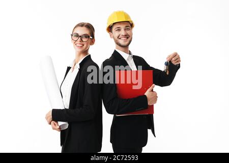 Young smiling business man in yellow safety helmet with folder and keys of new flat in hands, and woman in eyeglasses with plan happily looking in cam Stock Photo