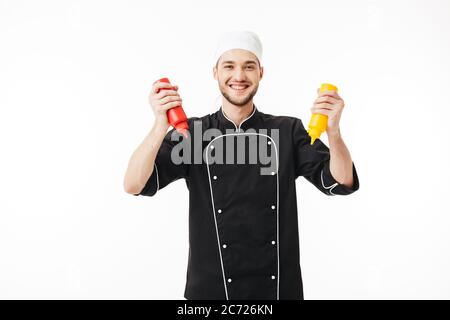 Young joyful male chef in black uniform holding bottles of mustard and ketchup in hands happily looking in camera over white background Stock Photo