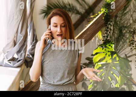 The surprised woman is unhappy with the phone conversation in her bright house. Angry girl speaks on the phone. Stock Photo