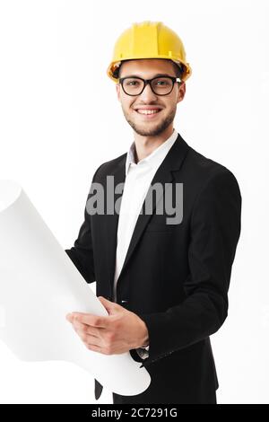 Young smiling bussinesman in eyeglasses and yellow safety helmet joyfully looking in camera holding sketch in hands over white background Stock Photo