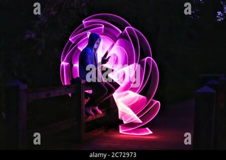 Boy sitting on a bridge with his smartphone in his hand. Violet light effect with a light saber in lightpainting. Stock Photo