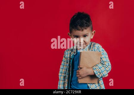 Photo of an adorable boy who looks into the camera, smiles and holds a book in his hands on a red background. Vintage tone Stock Photo