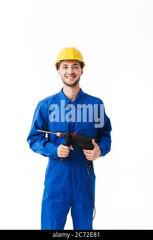 Young foreman in blue uniform and yellow helmet happily looking in camera holding drilling machine in hands over white background Stock Photo