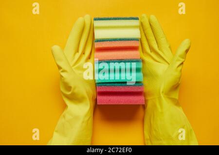 Close-up of woman in rubber gloves holding colorful sponges in a row isolated on yellow background Stock Photo