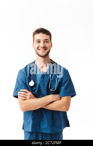 Young joyful male doctor in uniform with phonendoscope on neck happily looking in camera with cross hands over white background Stock Photo