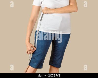 Caucasian mature adult woman dressed in a white T-shirt and a blue denim capris is going to shorten them and drawing a cut line on the desired length. Stock Photo