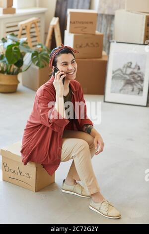Young smiling woman sitting on the box with things and talking on mobile phone during relocation Stock Photo