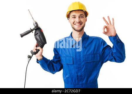 Young smiling foreman in blue uniform and yellow helmet joyfully showing ok gesture looking in camera holding drilling machine in hand over white back Stock Photo