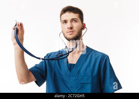 Young male doctor in uniform using phonendoscope thoughtfully looking aside over white background Stock Photo