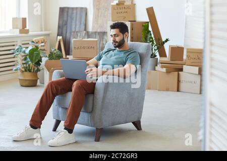 Young bearded man sitting on armchair and using laptop computer he moving to new apartment Stock Photo