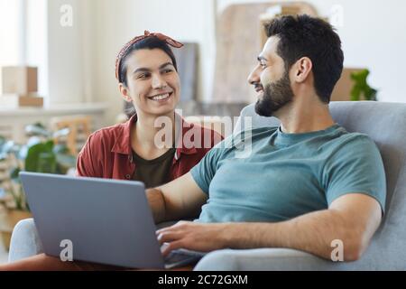 Young happy couple sitting on sofa talking to each other and using laptop while resting at home Stock Photo