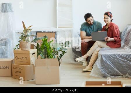 Young couple sitting on sofa among boxes with things and using laptop computer in new apartment Stock Photo