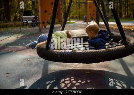 two toddlers a boy and a girl play cheerfully and swing on a swing hammock on a playground in the autumn park Stock Photo