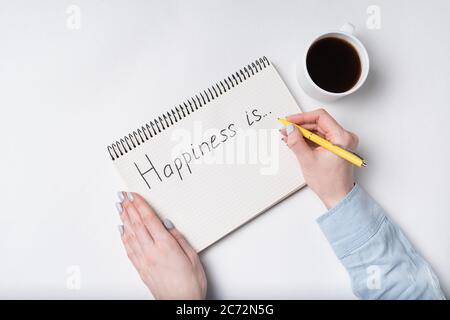 Copybook with HAPPINESS IS text. top view of female hands writing in Notebook and cup of coffee/ Stock Photo