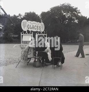 1950s, historical, a female ice-cream seller sitting outside with a lady customer at her mobile stall, beside the iron base of the famous Eiffel Tower in Paris, France. Stock Photo