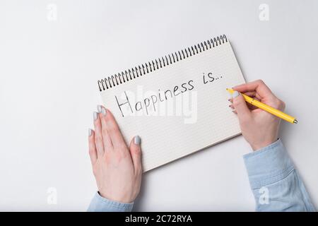 Copybook with HAPPINESS IS text. Top view of female hands writing in Notebook. Stock Photo