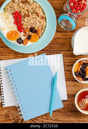 Notebook for diet plan and granola bowl with fruits on wooden background. Overhead. Stock Photo