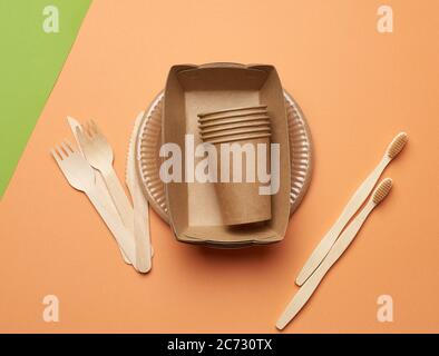 paper plates and cups from brown craft paper and wooden forks and knive on a orange background. Plastic rejection concept, zero waste Stock Photo