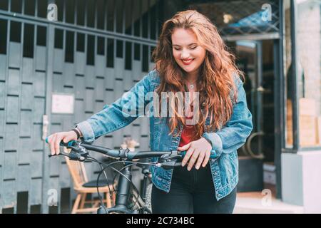 Red curled long hair caucasian teen girl on the city street walking with bicycle fashion portrait. Natural people beauty urban life concept image.
