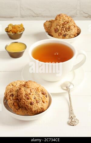 Oatmeal cookies with walnuts and raisins on small saucers and a cup of green tea, standing on a white wooden table against a white brick wall. Closeup Stock Photo