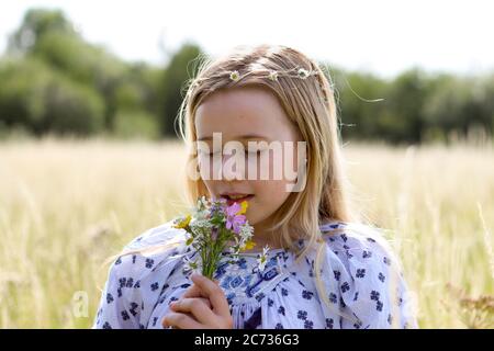 A young pretty hippy girl with daisy chain in her blonde hair holds a posy of wild flowers in the sunshine in a field of pasture in summertime. Stock Photo