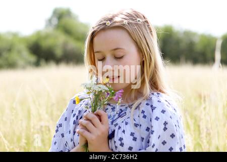 A young pretty hippy girl with daisy chain in her blonde hair holds a posy of wild flowers in the sunshine in a field of pasture in summertime. Stock Photo