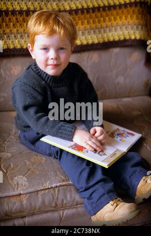 1990s RED HAIRED BOY ON COUCH WITH CHILDREN’S BOOK  =LOOKING AT CAMERA - kj13735 LGA001 HARS RED HAIR BABY BOY CHILDHOOD COMMUNICATE COOPERATION GROWTH JUVENILES YOUNGSTER CAUCASIAN ETHNICITY OLD FASHIONED Stock Photo